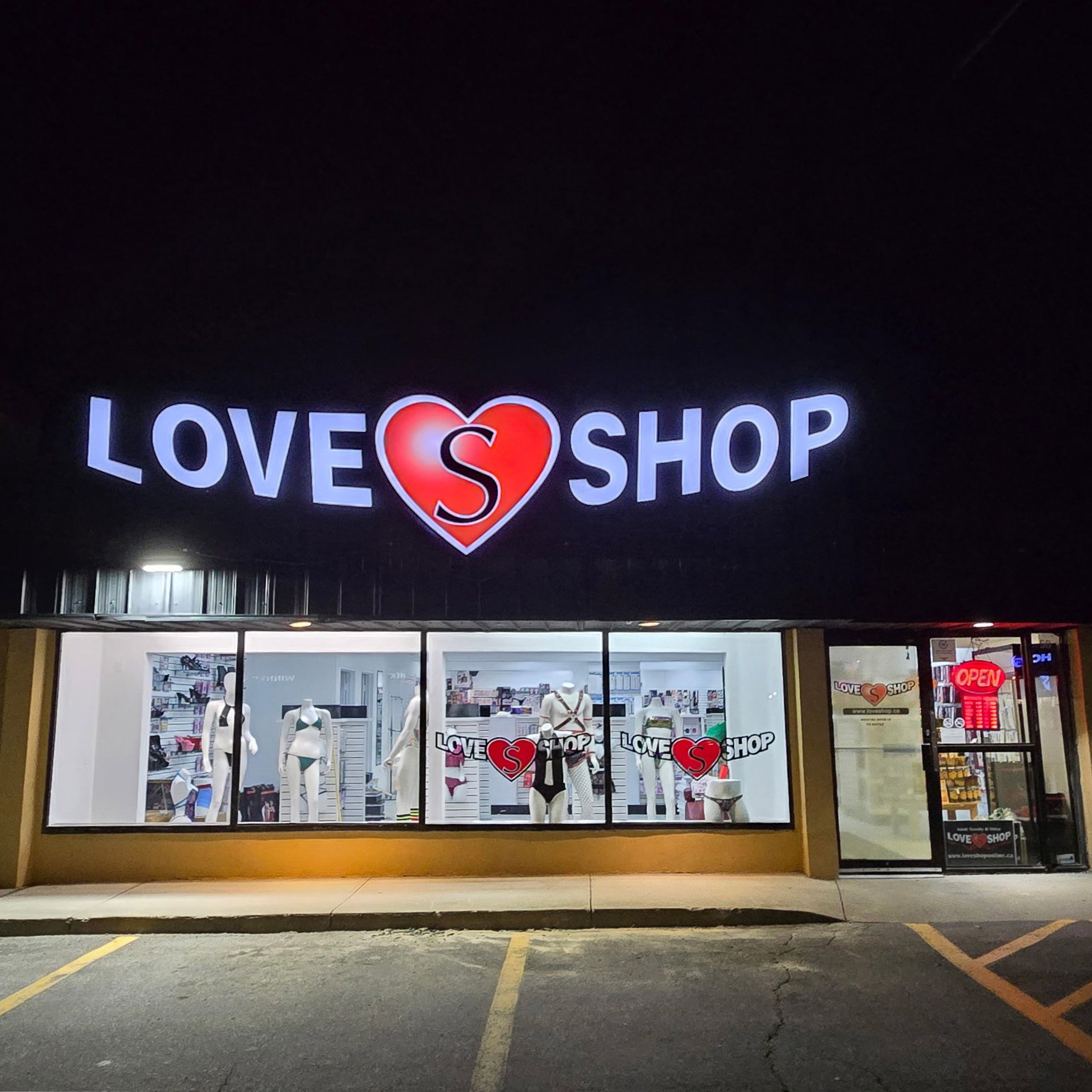 A storefront at night with a large illuminated sign reading LOVE SHOP, where the O is shaped like a red heart with a black script letter S inside. The shop windows display lingerie and other apparel, and a red heart sign is visible on the glass.